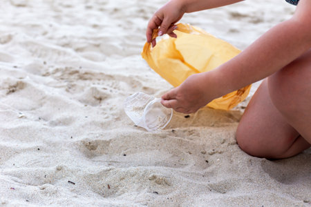 Individual is kneeling on sandy beach, carefully picking up litter with a yellow bag, showcasing commitment to environmental protection and cleanliness, encouraging others to maintain clean beachesの写真素材