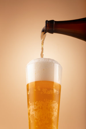 Golden beer pours from bottle into tall glass, creating thick foam and visible bubbles. Warm gradient background adds sense of refreshment and movement. Beer foam rising in tall glassの写真素材