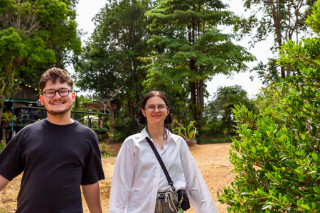 Two young adults outdoors in lush green environment, smiling and enjoying nature, with trees and pathway creating atmosphere of friendship and connection. Young adults together in natural settingの写真素材