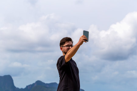 Young man with glasses is taking selfie outdoors against backdrop of mountains and cloudy sky, capturing moment of joy and connection with nature. Male individual is taking selfieの写真素材