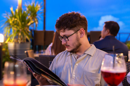 Young man with glasses is focused on reading menu at rooftop restaurant, surrounded by soft lighting...