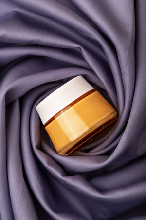 Jar of face cream. Yellow cosmetic cream jar is elegantly positioned on soft purple satin fabric, showcasing its smooth surface and rich color, surrounded by gentle foldsの写真素材