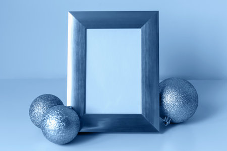 Mockup. Christmas. Empty silver frame positioned beside three glittering blue ornaments on soft blue...
