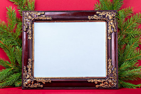 Mockup. Christmas. Ornate wooden picture frame with intricate detailing placed among lush evergreen branches, creating festive atmosphere against bright red background, perfect for seasonal displaysの写真素材