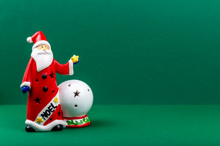 Christmas. Santa Claus. Festive Santa Claus figurine wearing red coat stands beside snow globe on green background, creating cheerful atmosphere for holiday decoration and seasonal celebrationsの写真素材