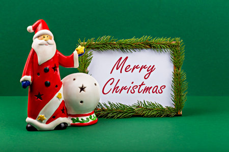 Merry Christmas. Santa Claus. Festive Santa Claus figurine in vibrant red attire stands next to decorative sign that wishes Merry Christmas, framed with lush greenery against green backgroundの写真素材