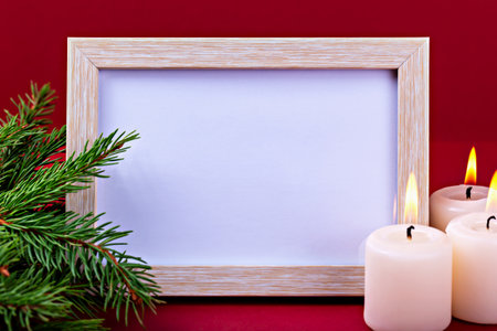Christmas. New Year. Empty wooden frame with copy space is placed on red surface, surrounded by lit candles and fresh evergreen branches, creating warm and inviting atmosphere for holiday celebrationsの写真素材