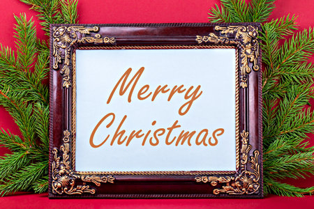 Merry Christmas. Elegant wooden frame displays festive message, surrounded by vibrant green pine branches against rich red background, creating warm and inviting holiday atmosphereの写真素材