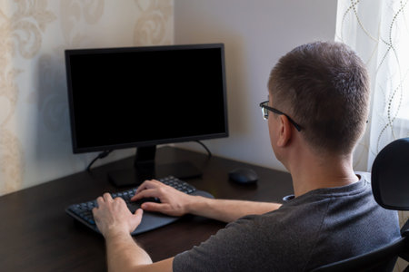 Remote work from home. Man with short hair is focused on typing at computer in modern workspace, featuring sleek desk, keyboard and soft natural light filtering through windowの写真素材