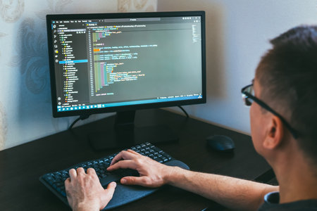 Remote work from home. Male programmer is engaged in coding on a computer, showcasing modern workspace with sleek monitor and keyboard, emphasizing concentration and digital environmentの写真素材