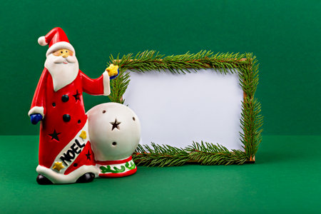Christmas. Mockup. Santa Claus. Festive Santa figurine with red attire stands next to decorative snow globe, all set against vibrant green background, featuring empty frame adorned with pine branchesの写真素材