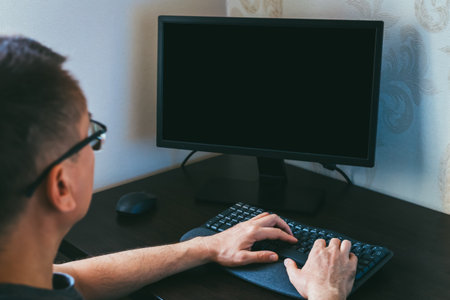 Remote work from home. Male individual wearing glasses is focused on typing on keyboard in contemporary workspace, with blank computer screen and minimalistic decor creating atmosphere of productivityの写真素材