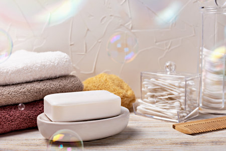 Health and hygiene. White soap bar placed on dish, surrounded by neatly folded towels and bathroom accessories, with colorful bubbles floating in air, creating serene and clean atmosphereの写真素材