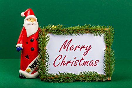 Merry Christmas. Santa Claus. Festive Santa Claus figurine in red attire stands next to decorative frame adorned with green foliage, displaying cheerful holiday greeting, creating joyful atmosphereの写真素材