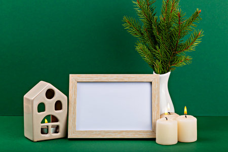 Mockup. Christmas. Decorative arrangement featuring an empty wooden frame, three lit candles, ceramic house, and an evergreen plant, creating cozy ambiance against vibrant green backgroundの写真素材