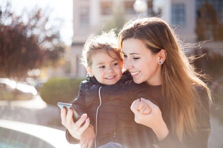 Young smiling woman and little happy girl reading message on smartphone. Autumn city backgroundの写真素材