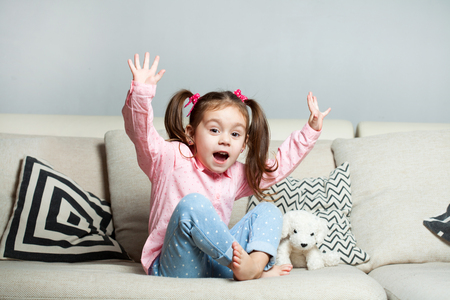 Pretty happy little girl in casual wearing sitting on sofa with toy dog and smiling.の写真素材