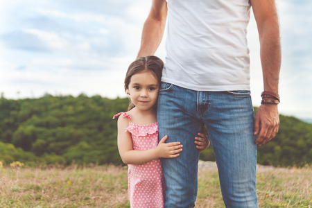 Pretty smiling little girl hugging father's leg. Summer happy family day in forestの写真素材