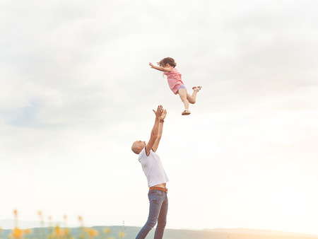 Happy Father playing with his daughter in sunny field on sunset. Summer happy family lifestyle, truly emotions. Little girl flying high in the sky from dad's handsの写真素材