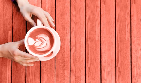 Woman's hands holding cup of cappuccino on wooden table. Living Coral colored picture. Beverage...