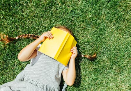 Cute schoolgirl with pigtails laying on grass while reading book. Education, learning and back to school concept.の写真素材
