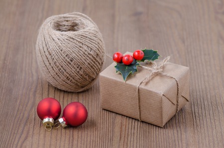 a gift box with twine ribbon and holly berries and two red baubles on brown wooden backgroundの写真素材