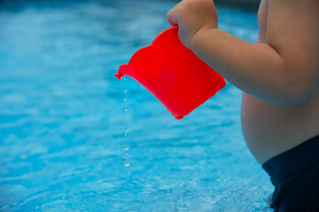 Child's hand holding a red plastic watering can in swimming pool.の写真素材