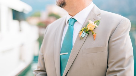 The groom with a beard boutonniere on the lapel of a light jacket with a blue tie.
