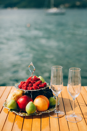 Two-storeyed plate with fruits: raspberries, peach, apples, figs, pears, grapes. And two glasses for champagne on the table near the sea.の写真素材