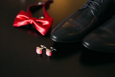 Black shoes of the groom, red bow tie, cufflinks, belt, on a black background with bright bokeh. Wedding groom accessories.の写真素材