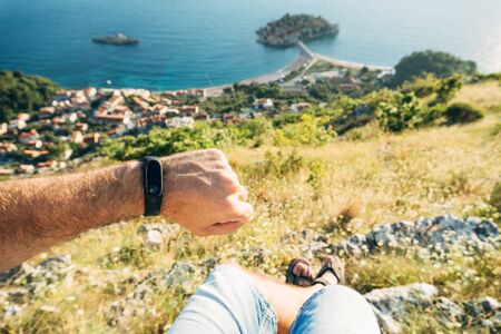 Fitness bracelet on a man's hand. A man sits on top of a mountain overlooking the island of Sveti Stefan in Montenegroの写真素材