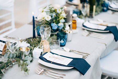 A square plate with a blue cloth towel, knives and forks next to the plate. Flower composition with eucalyptus leaves in the center of the table and burning candles.の写真素材