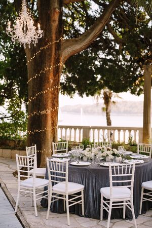 Wedding dinner table reception. Round banquet tables for guests with gray tablecloth. Floral arrangement of roses in center of the table. Garland on tree trunks, crystal chandeliers hang in airの写真素材