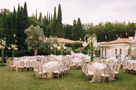 Wedding dinner table reception. Elegant tables for guests with cream tablecloths with patterns, on green lawn, with garlands and chandeliers hanging over them. Chairs with round backの写真素材