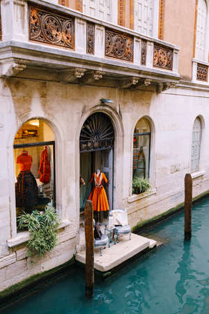 Venice, Italy - 04 october 2019: Showcase of a fashion clothing store in Venice, Italy. Stylish orange dress 60-70s on a mannequin. The threshold of the store in azure water.のeditorial素材