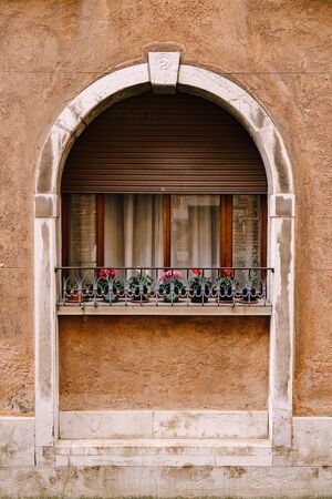 Close-ups of building facades in Venice, Italy. A stone arch above a wooden window on the facade of building. White curtains in window, small flowers in pots on windowsill with a metal forged fenceの写真素材