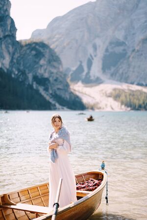 A bride stands in a wooden boat at the Lago di Braies in Italy. Wedding in Europe, on Braies lake. A young girl in a white wedding dress, covers her shoulders with a blue sweater.の写真素材