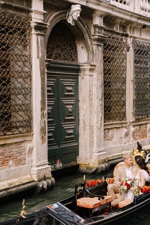 Italy wedding in Venice. Gondolier rolls bride and groom in classic wooden gondola along narrow Venetian canal. Newlyweds are sitting in boat nose to nose, swim against backdrop of old forged latticeの写真素材