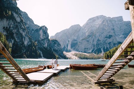 The bride and groom walk on a wooden boat dock at Lago di Braies in Italy. Wedding in Europe, at Braies lake. Newlyweds walk, kiss, cuddle against the backdrop of rocky mountains.の写真素材