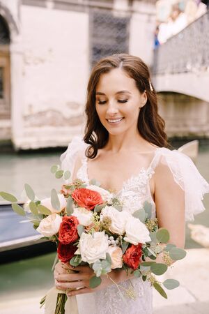 Venice wedding in Italy. A bride in a white dress, with a train, with a bouquet of white and red roses in her hands, stands on the pier near the moored gondola in a narrow Venetian canal.の写真素材