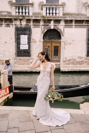 Venice wedding in Italy. A bride in a white dress, with a train, with a bouquet of white and red roses in her hands, stands on the pier near the moored gondola in a narrow Venetian canal.の写真素材