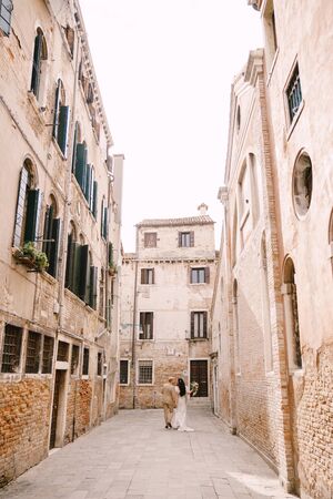 Italy wedding in Venice. The bride and groom walk along the deserted streets of the city. Newlyweds are walking in a dead end alley on the background of brick buildings.の写真素材