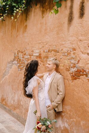 Italy wedding in Venice. The bride and groom walk along the deserted streets of the city. Newlyweds are standing nearby and hugging near the orange wall of a beautiful house.の写真素材