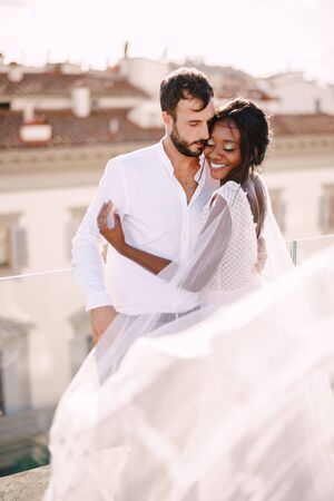 Destination fine-art wedding in Florence, Italy. Multiethnic wedding couple. African-American bride hugs Caucasian groom on the roof with cityscape view.の写真素材
