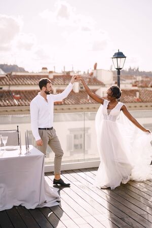 Interracial wedding couple. Destination fine-art wedding in Florence, Italy. African-American bride and Caucasian groom are dancing near the table for a wedding dinner, on the roof of the restaurant.の写真素材