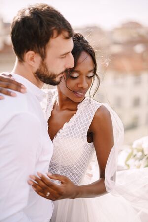 Destination fine-art wedding in Florence, Italy. Interracial wedding couple. Caucasian groom and African-American bride cuddling on a rooftop in sunset sunlight.の写真素材