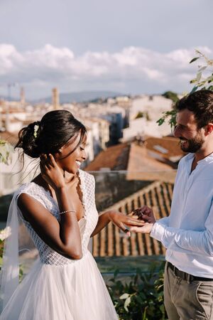 Multiethnic wedding couple - African-American bride and Caucasian groom. Groom puts a ring on brides finger. Destination fine-art wedding in Florence, Italy. Wedding ceremony on the roofの写真素材