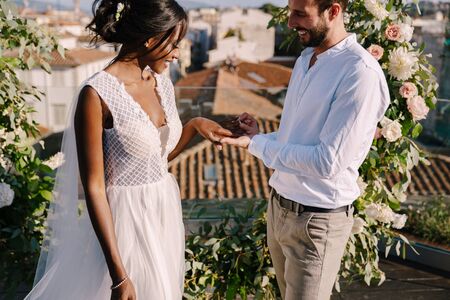 Mixed-race wedding couple - African-American bride and Caucasian groom. Groom puts a ring on brides finger. Destination fine-art wedding in Florence, Italy. Wedding ceremony on the roofの写真素材