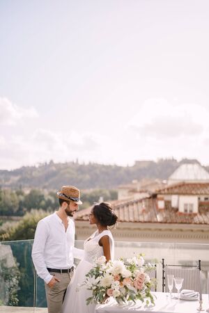 Destination fine-art wedding in Florence, Italy. Multiethnic wedding couple. African-American bride and Caucasian groom with a beard and straw hat, near the table for a wedding dinner.の写真素材