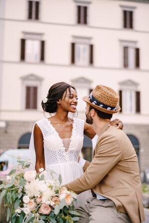 Multiracial wedding couple. Wedding in Florence, Italy. African-American bride in a white dress with a long veil and a bouquet, and Caucasian groom in a sandy jacket and straw hat.の写真素材
